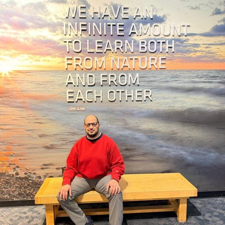 Ensar Seker sitting on a bench beneath a John Glenn quote about learning from nature and from each other